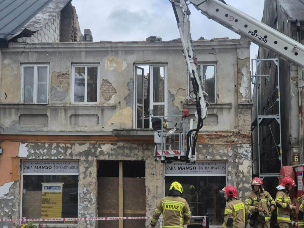 Grupa strażaków w hełmach pracuje przy zniszczonym budynku, obok podnośnika i rusztowania.