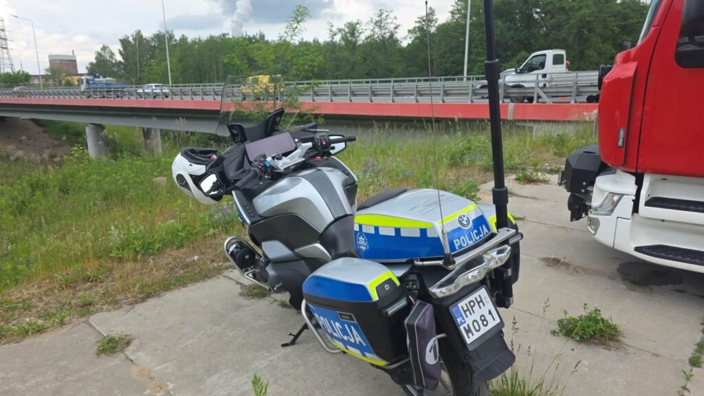 Policyjny motocykl zaparkowany na poboczu drogi, w tle most oraz przejeżdżające pojazdy.