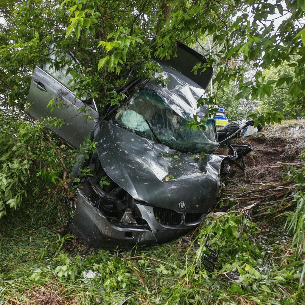 Wrak czarnego samochodu osobowego wjeżdżającego w krzewy, z uszkodzoną przednią częścią i rozbitą szybą.