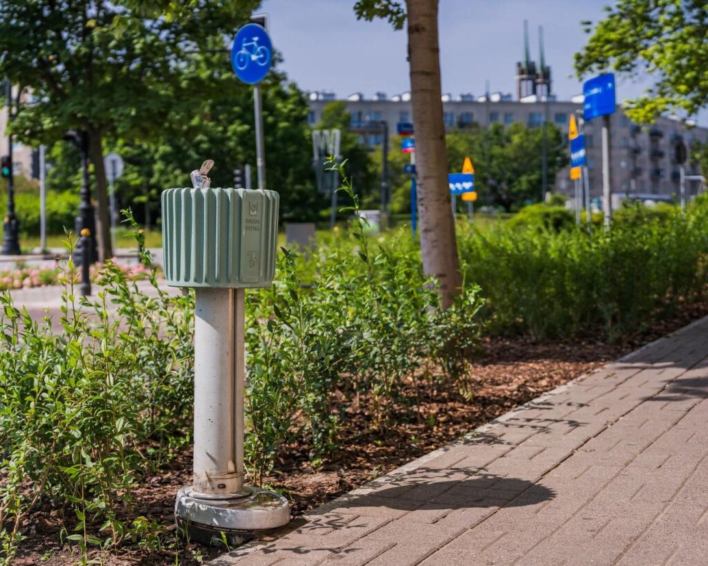 Zielony hydrant w miejskim otoczeniu, otoczony niską roślinnością i chodnikiem, na tle budynków i znaków drogowych.