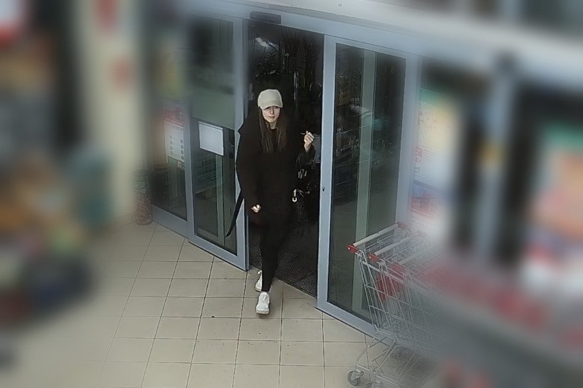 Kobieta w czarnej kurtce i beżowej czapce wychodzi ze sklepu, trzymając torbę na zakupy. W tle widać wózek sklepowy.