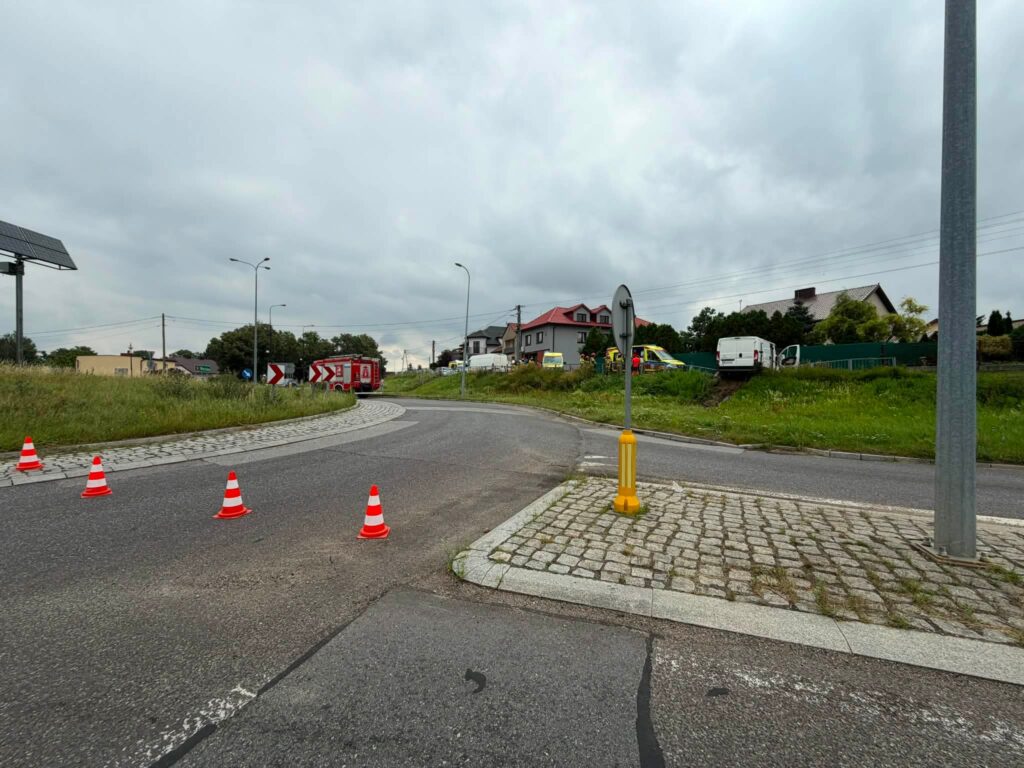 Na rondzie ustawione są pachołki drogowe, a w tle widoczne pojazdy służb ratunkowych oraz budynki mieszkalne.