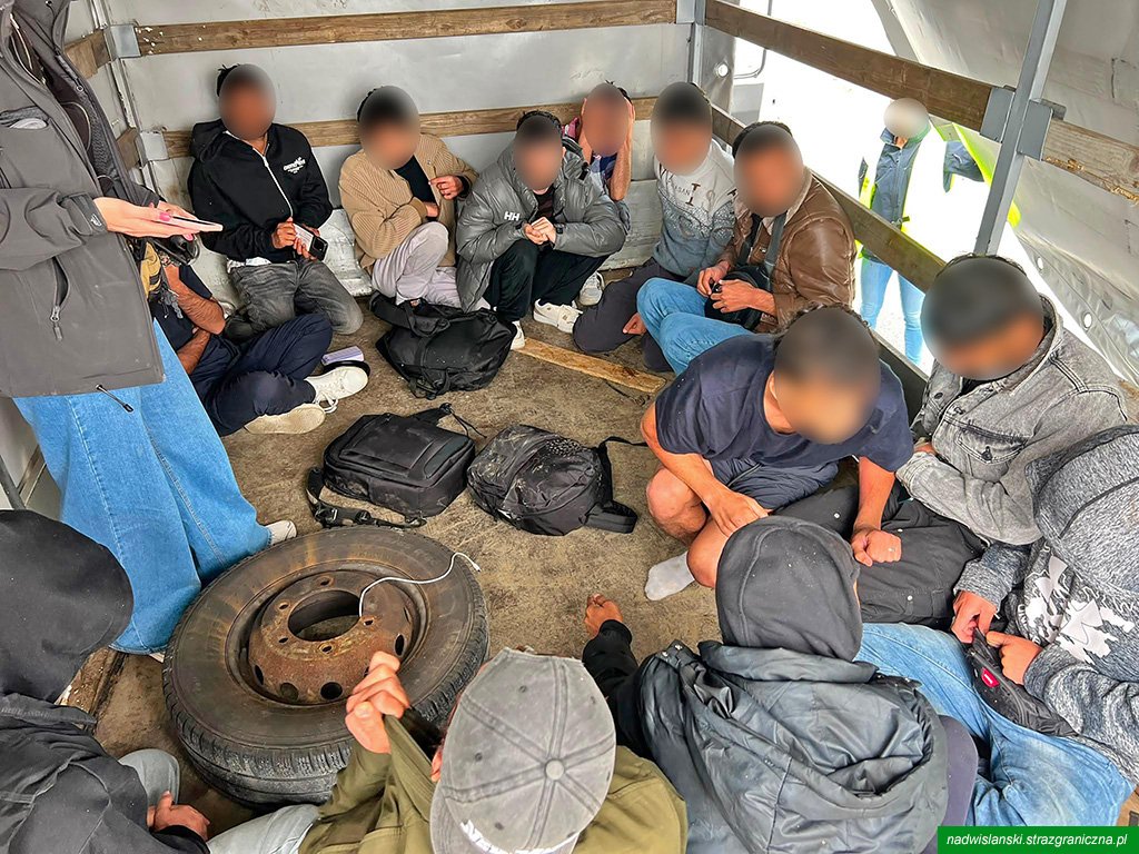 Grupa osób siedzących w zamkniętej przestrzeni, otoczona drewnianymi ściankami, z torbami i kołem samochodowym na podłodze.