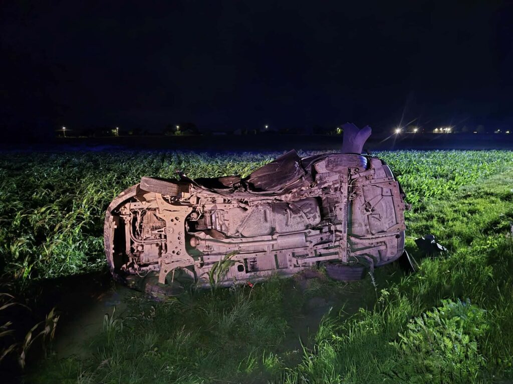 Odwrócone auto leży na polu, oświetlone przez reflektory, otoczone wysoką trawą w nocy.