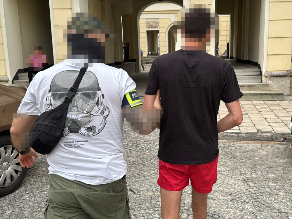 Policjant prowadzi zatrzymanego mężczyznę w szortach w otoczeniu architektury miejskiej.