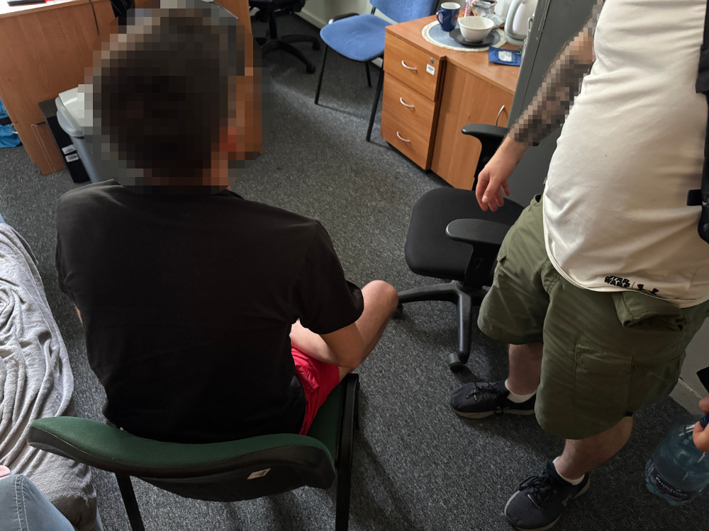 Mężczyzna w czarnej koszulce siedzący na krześle, obok niego stoi drugi mężczyzna w białej koszulce, w biurowym wnętrzu.