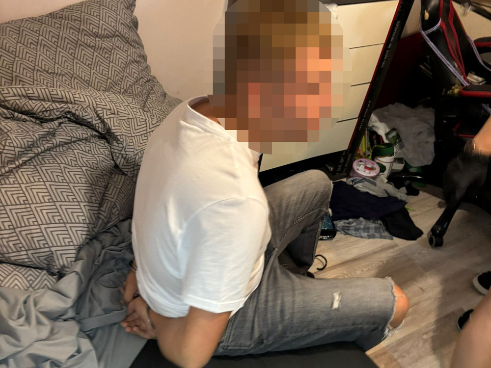 Mężczyzna w białej koszulce siedzi na podłodze z rękami związanymi z tyłu, w nieuporządkowanym pokoju.