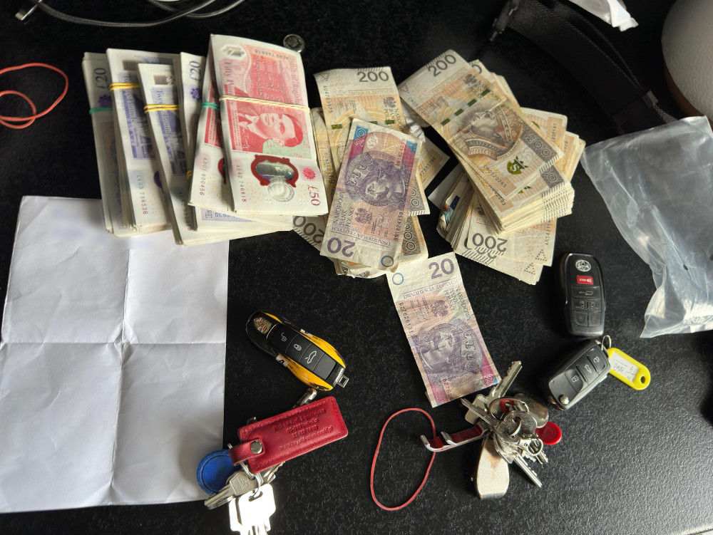 Na czarnym stole leżą stosy banknotów w różnych nominałach, kluczyki do samochodów oraz złożona kartka papieru.