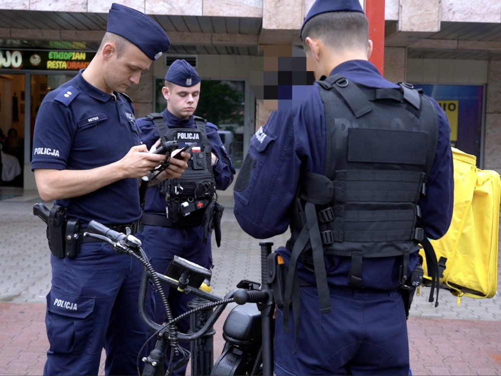 Trzech policjantów w mundurach kontroluje elektryczne jednoślady w miejskim otoczeniu.