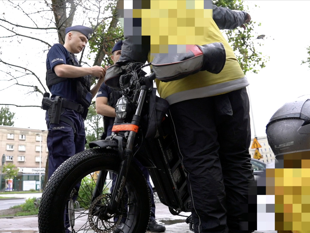 Dwaj policjanci kontrolują elektryczny jednoślad na chodniku, podczas gdy operator pojazdu w żółtej kamizelce odpowiada na pytania.