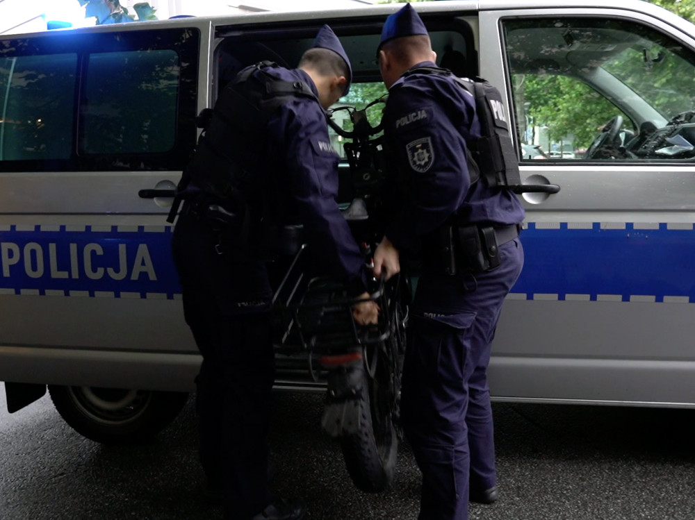 Dwaj policjanci w mundurach umieszczają elektryczny jednoślad do radiowozu na ulicy.