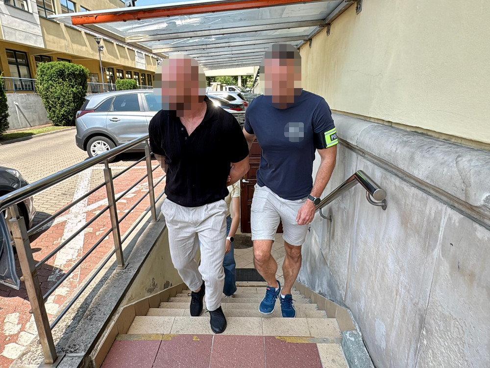 Mężczyzna w czarnej koszuli prowadzony przez policjanta w niebieskiej koszulce schodzi po schodach w otoczeniu samochodów.