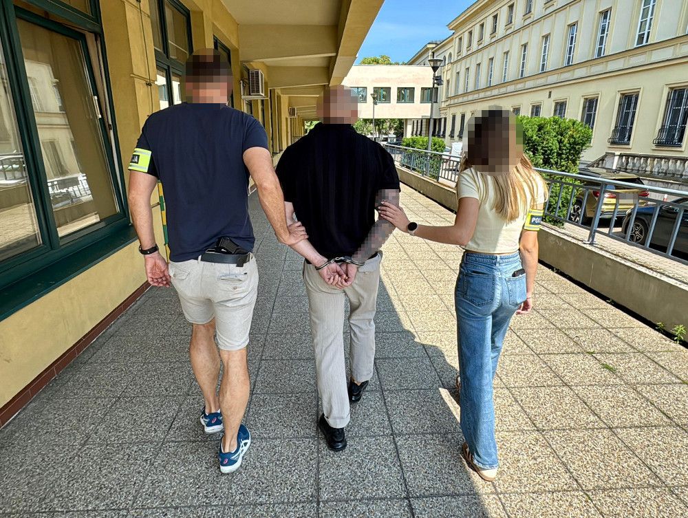 Mężczyzna w kajdankach prowadzony przez dwóch funkcjonariuszy policji w korytarzu budynku.