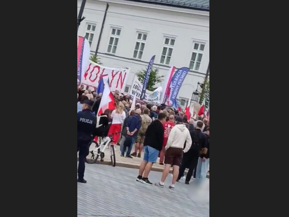 Tłum ludzi z flagami i transparentami gromadzi się na placu, w tle widoczny biały budynek. Policjant na rowerze obserwuje sytuację.