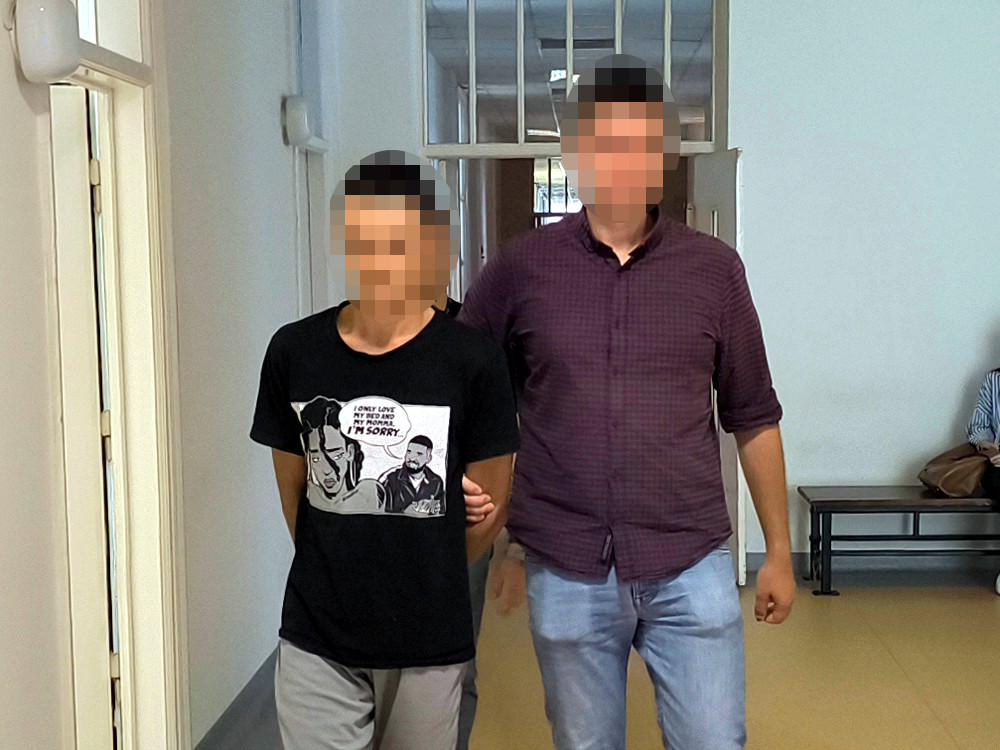Młody mężczyzna w czarnym t-shircie prowadzi przez policjanta w korytarzu budynku, na ścianie widać drzwi.