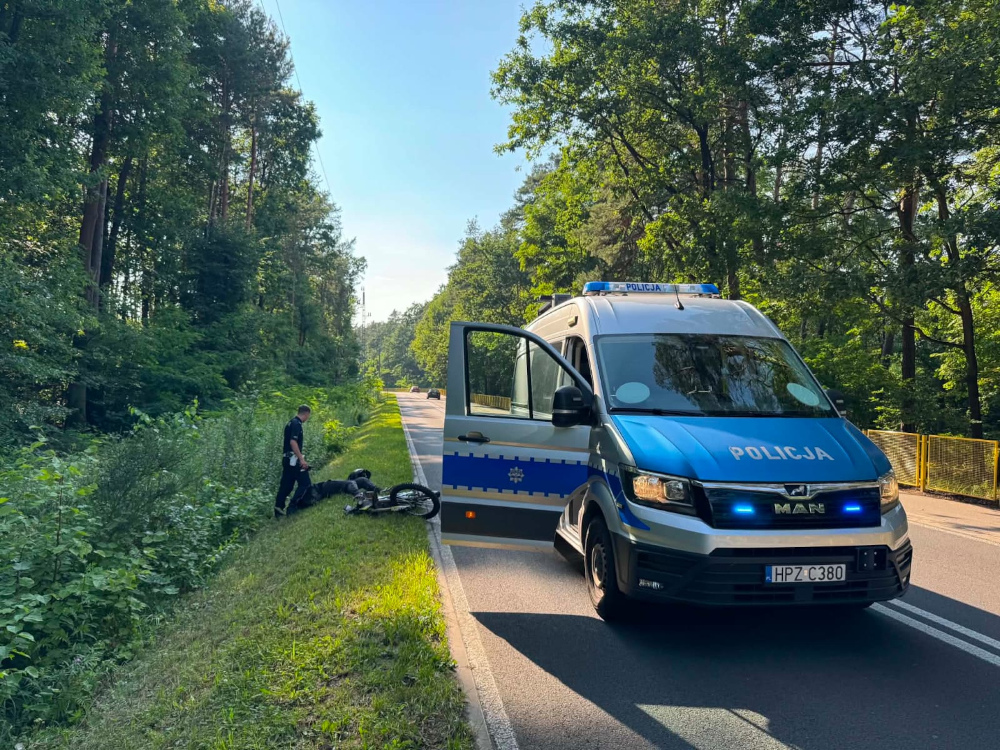 Policjant bada rower leżący na poboczu drogi w lesie, obok zaparkowanego radiowozu z włączonymi sygnałami.