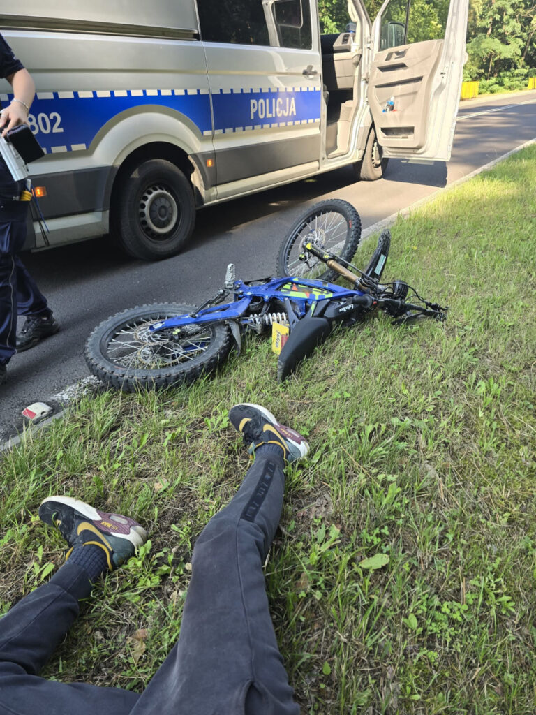 Mężczyzna w czarnych spodniach leży na trawie obok przewróconego motocykla, a w tle stoi radiowóz policji.
