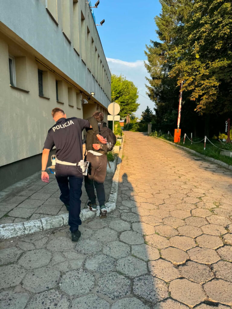 Policjant prowadzi młodego mężczyznę w stronę budynku, w tle widać zieleń drzew i asfaltową ścieżkę.