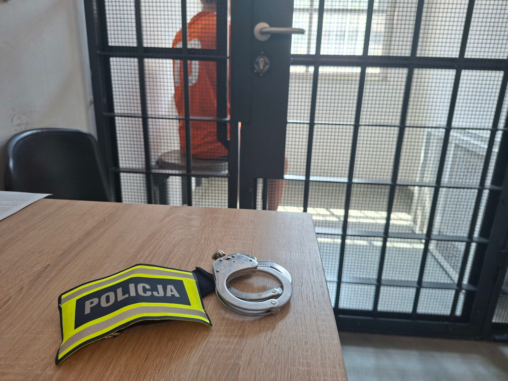 Mężczyzna w pomarańczowym stroju siedzi na krześle za kratami, obok leżą kajdanki i oznaczenie "POLICJA" na stole.