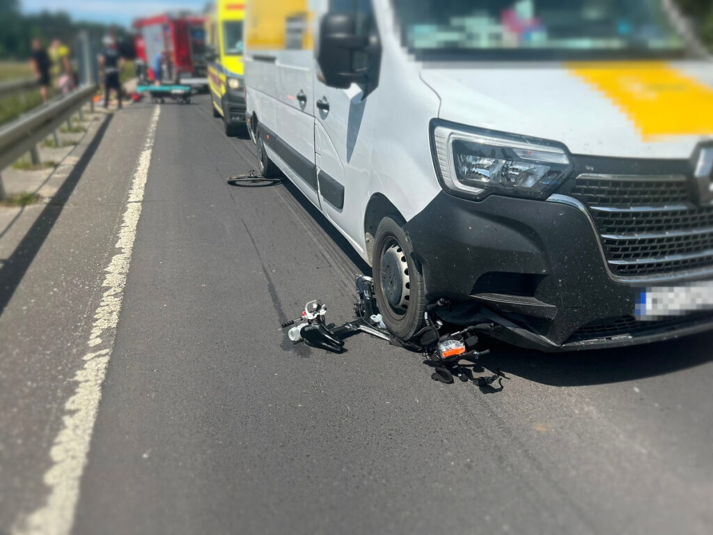 Rowerzysta leży na jezdni obok zniszczonego roweru, a obok stoi dostawcza furgonetka z uszkodzoną przednią częścią.