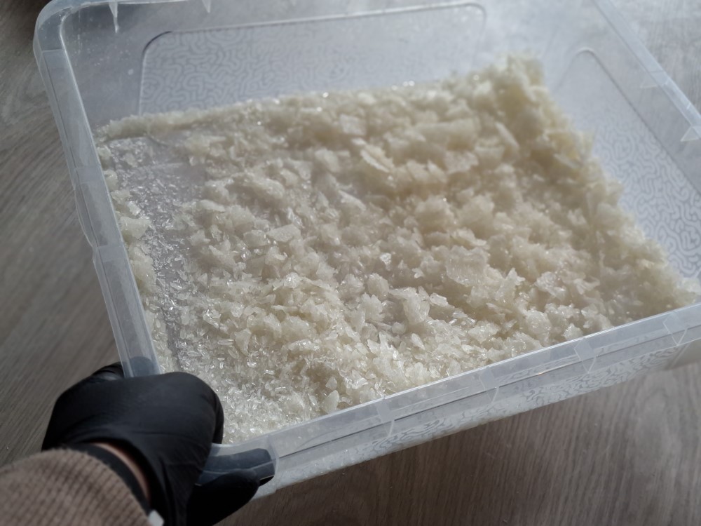 Ręka w rękawiczce trzyma przezroczysty pojemnik z dużą ilością kryształków substancji chemicznej.