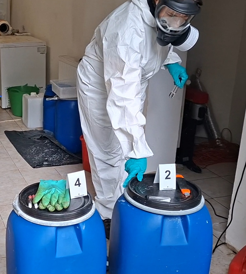 Funkcjonariusz w białym kombinezonie ochronnym otwiera niebieskie beczki, oznaczone numerami, w laboratorium.