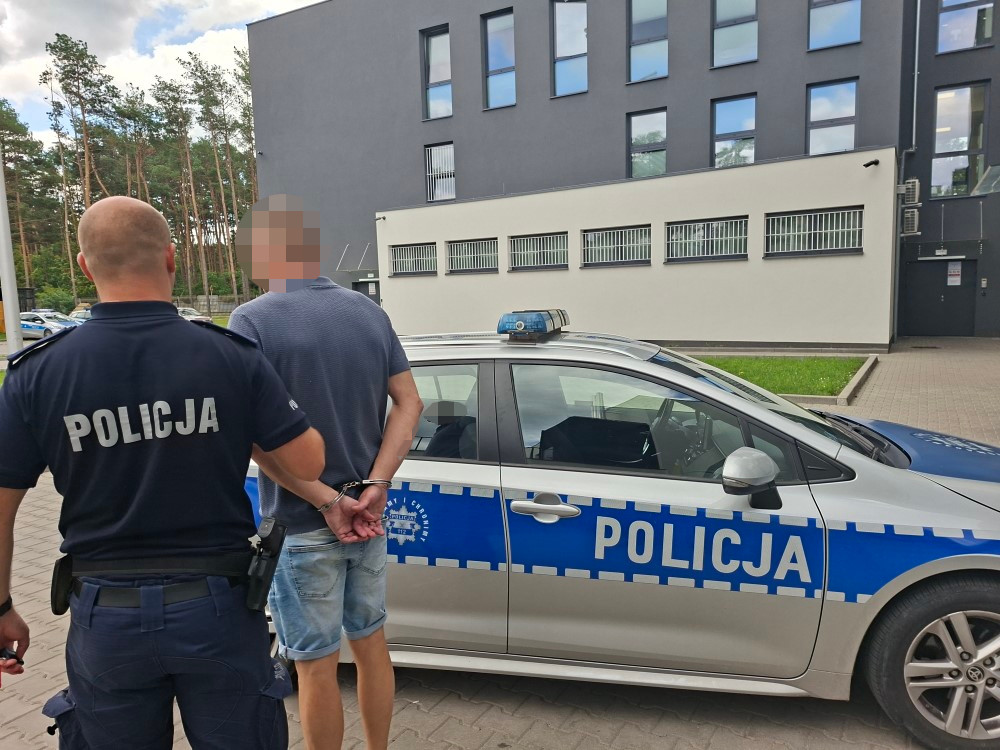 Policjant w mundurze prowadzi zatrzymanego mężczyznę w okolicy nowoczesnego budynku.