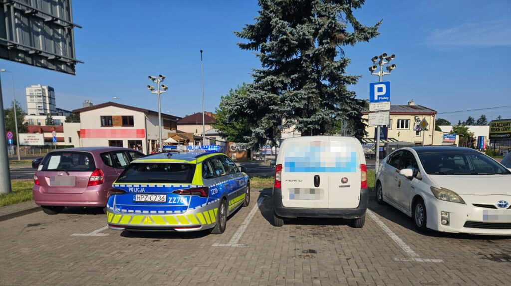 Radiowóz policyjny stoi na parkingu obok dwóch osobowych samochodów, w tle widoczny budynek i drzewa.