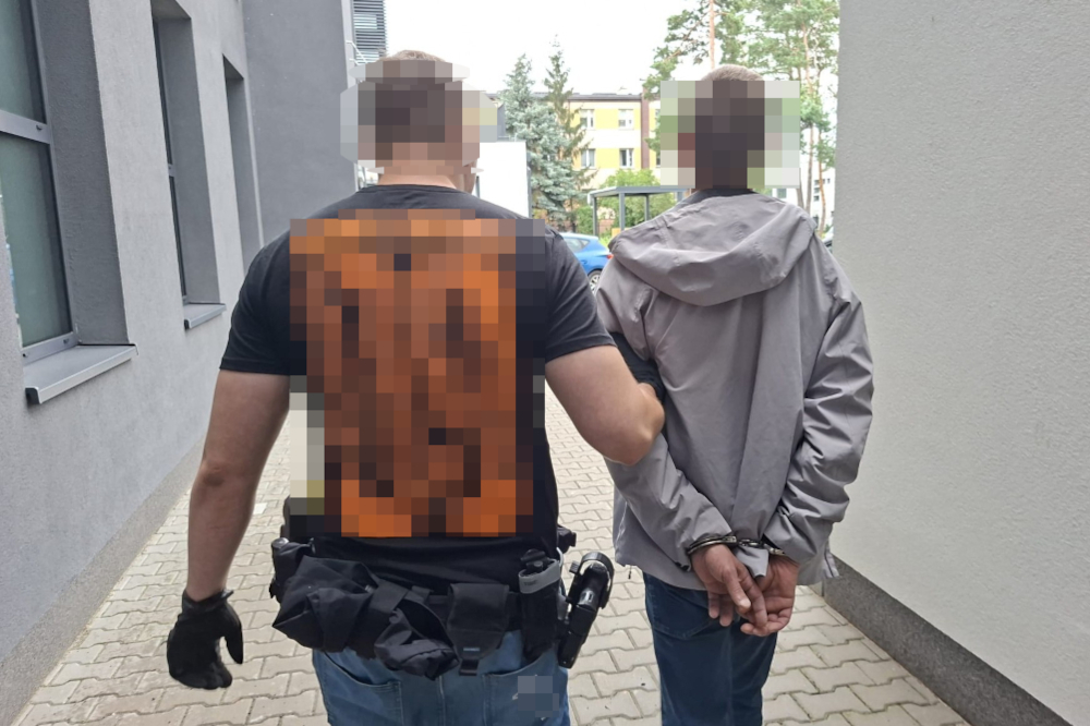 Mundurowy funkcjonariusz prowadzi aresztowanego mężczyznę z rękami związanymi za plecami po chodniku w miejskim otoczeniu.