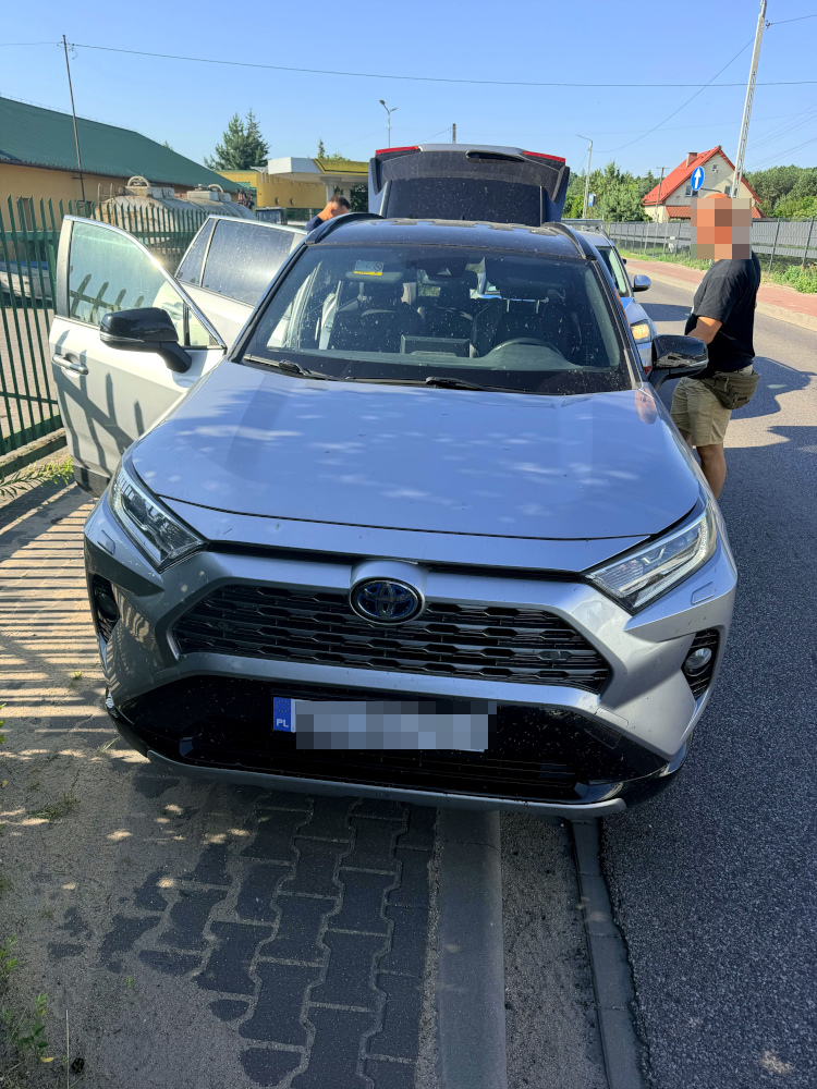 Kierowca w krótkich spodniach przeszukuje wnętrze skradzionej Toyoty na poboczu drogi w słoneczny dzień.