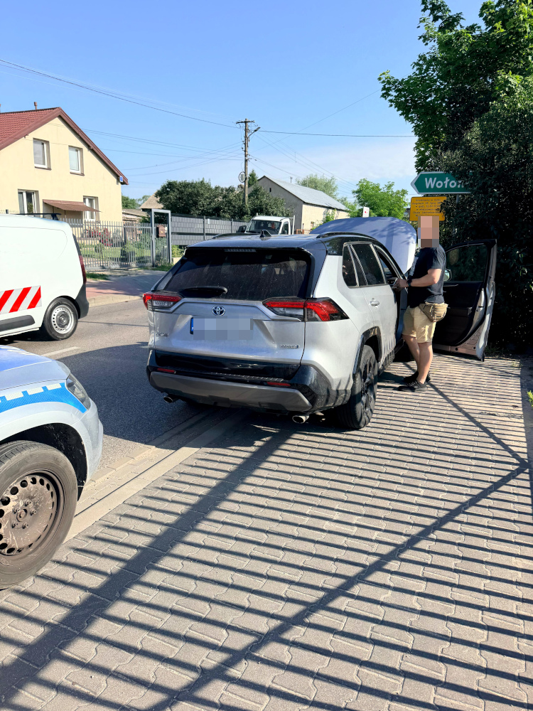 Mężczyzna w krótkich spodniach rozmawia obok zaparkowanego samochodu SUV na ulicy, w tle widoczny biały furgon.