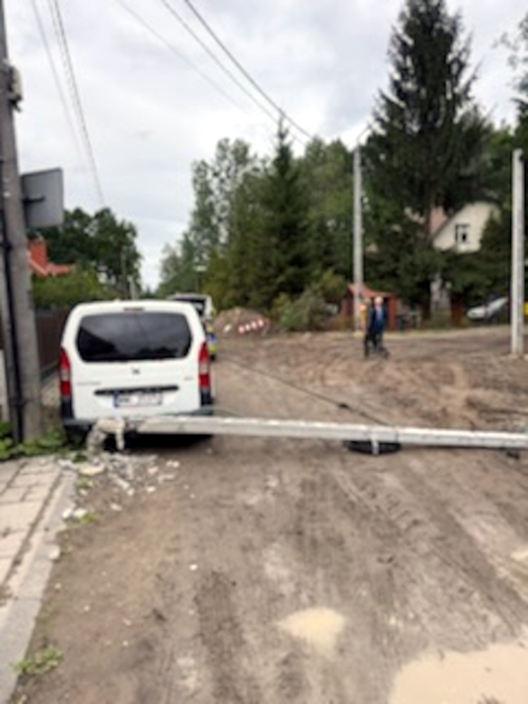 Biały van zaparkowany na drodze, z przewróconym słupem energetycznym leżącym na ziemi. W tle widać ludzi i drzewa.