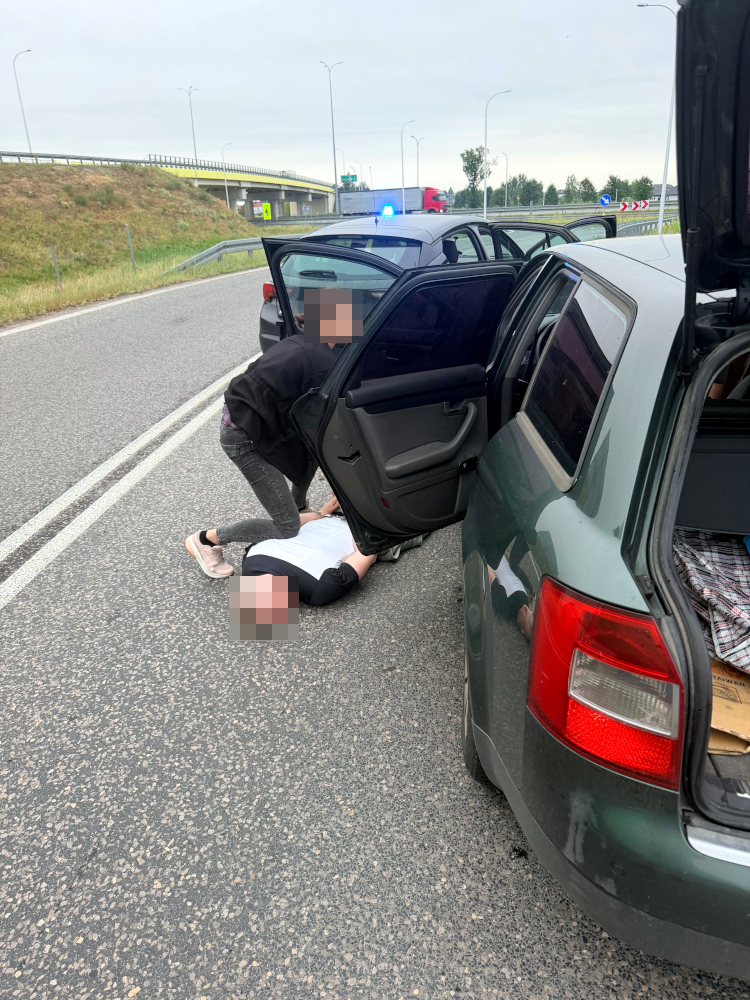 Kobieta leży na drodze, a mężczyzna w czarnej odzieży udziela jej pomocy, obok stoją samochody.