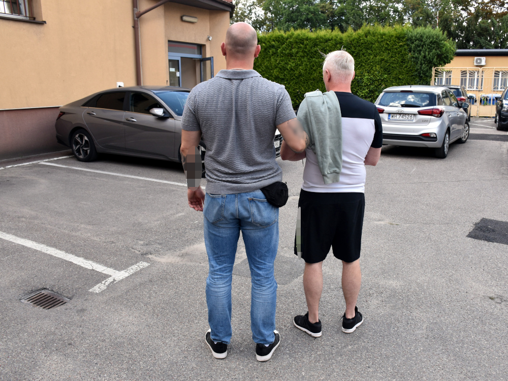 Mężczyzna w szarej koszulce prowadzi starszego mężczyznę w białej bluzie i szortach na parkingu przed budynkiem.