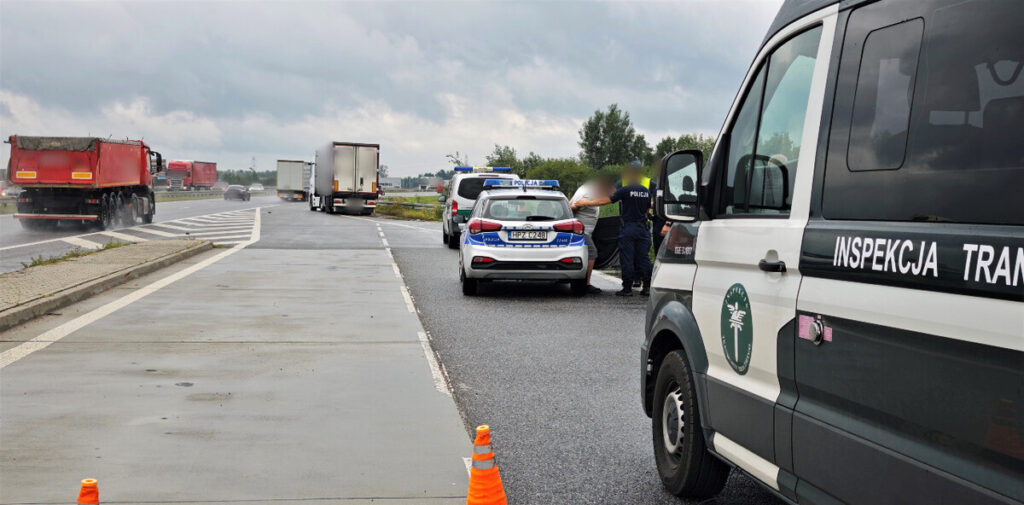 Funkcjonariusze policji sprawdzają dokumenty kierowcy przy drodze, obok zaparkowanego pojazdu Inspekcji Transportu Drogowego.