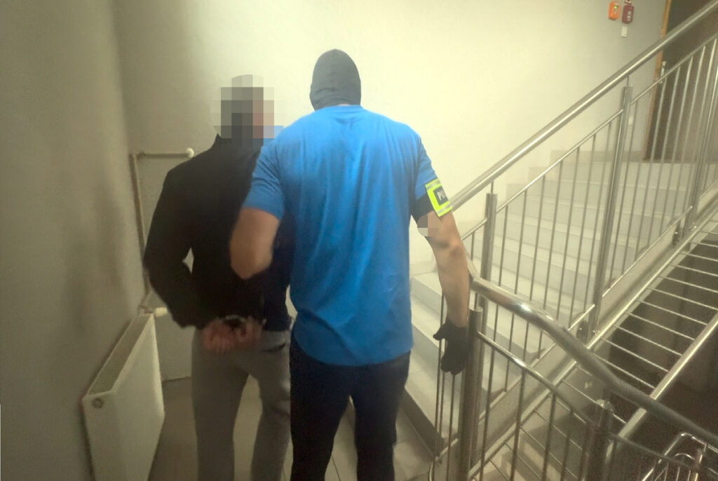 Mężczyzna w niebieskiej koszulce z oznaczeniem policji prowadzi zatrzymanego na klatce schodowej.