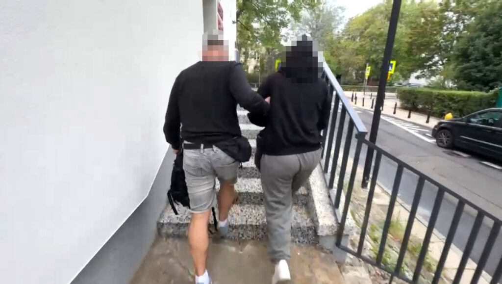 Mężczyzna prowadzi kobietę w szarej dresowej odzieży po schodach w otoczeniu zieleni i ulicy.