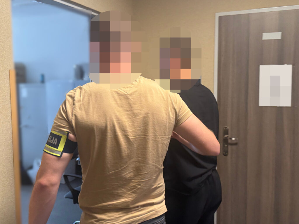 Mężczyzna z opaską na ramieniu rozmawia z drugą osobą w biurowym korytarzu.