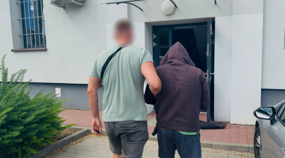 Mężczyzna w szarej koszulce prowadzi osobę w ciemnej bluzie w kierunku budynku z drzwiami wejściowymi.