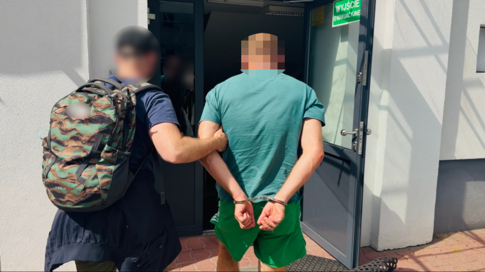 Mężczyzna w zielonej koszulce i szortach, z rękami skuty w kajdankach, prowadzony przez funkcjonariusza w kierunku drzwi.