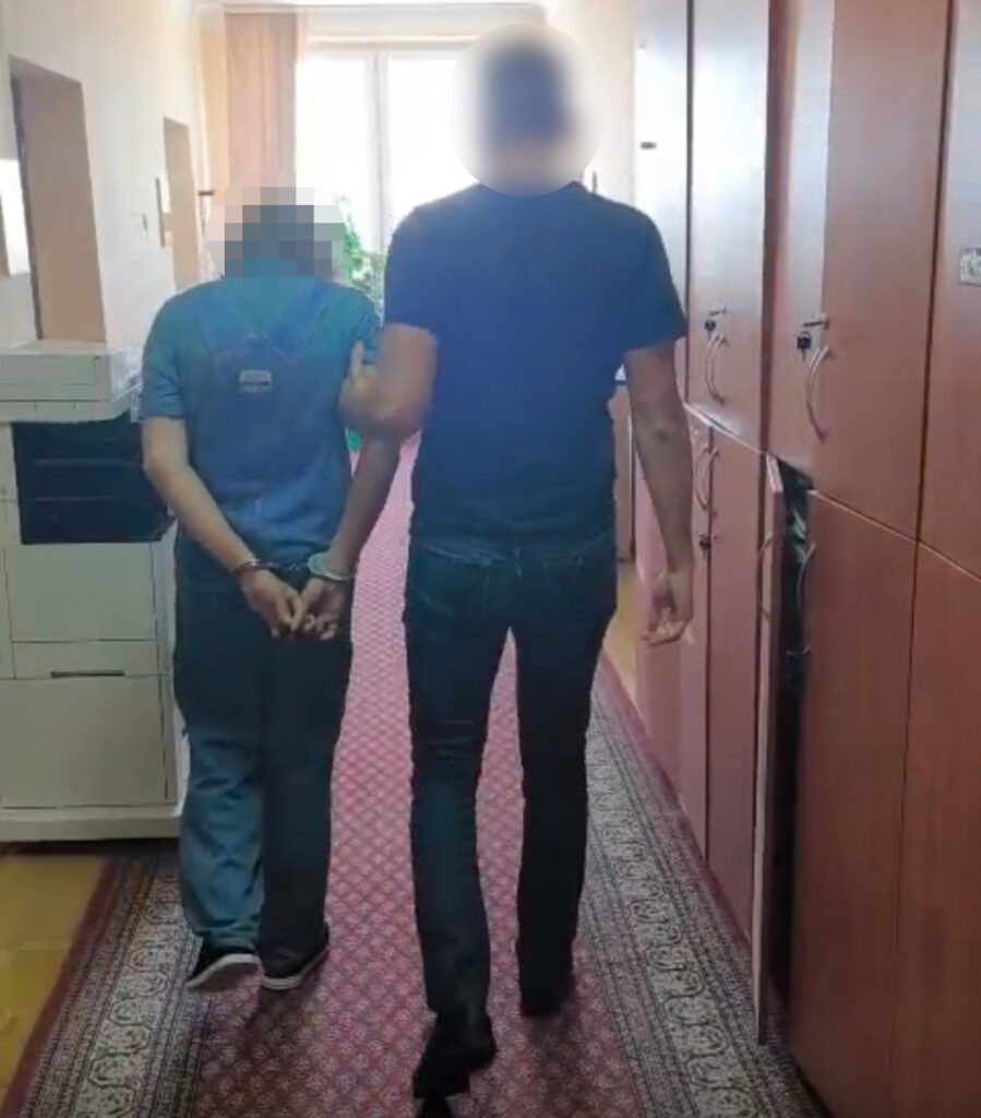 Mężczyzna w kajdankach prowadzony przez funkcjonariusza policji w korytarzu budynku.