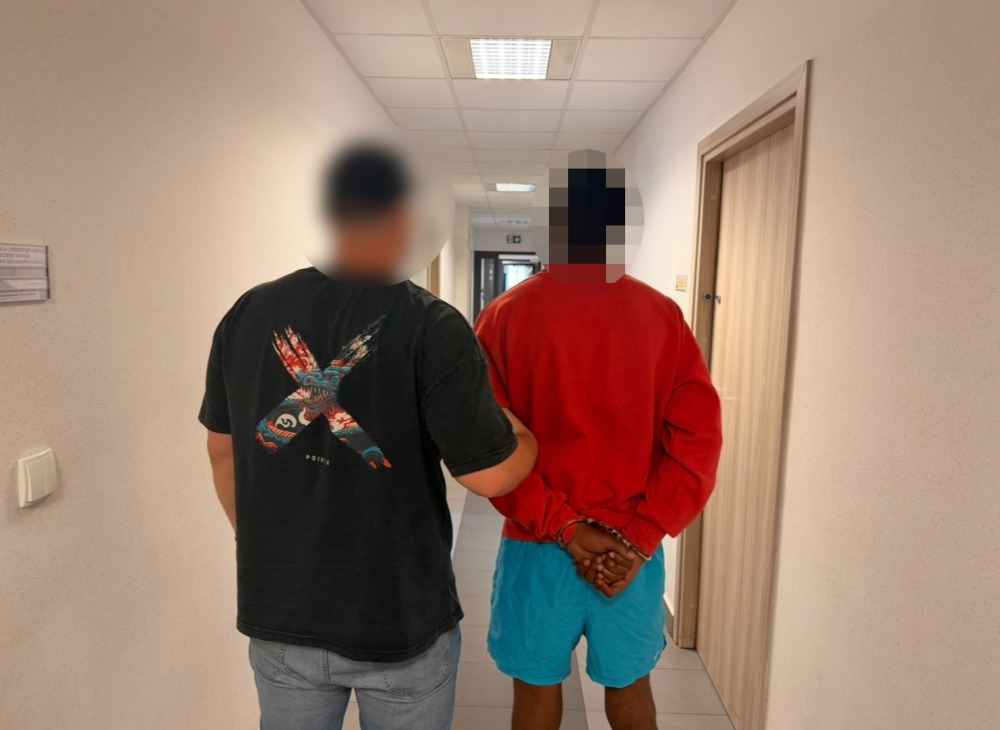 Mężczyzna w czerwonej bluzie i niebieskich szortach prowadzi przez funkcjonariusza w korytarzu budynku.