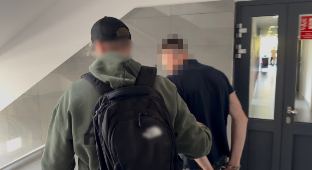 Mężczyzna w zielonej bluzie z kapturem i plecakiem idzie korytarzem, obok niego drugi mężczyzna w ciemnej koszulce.
