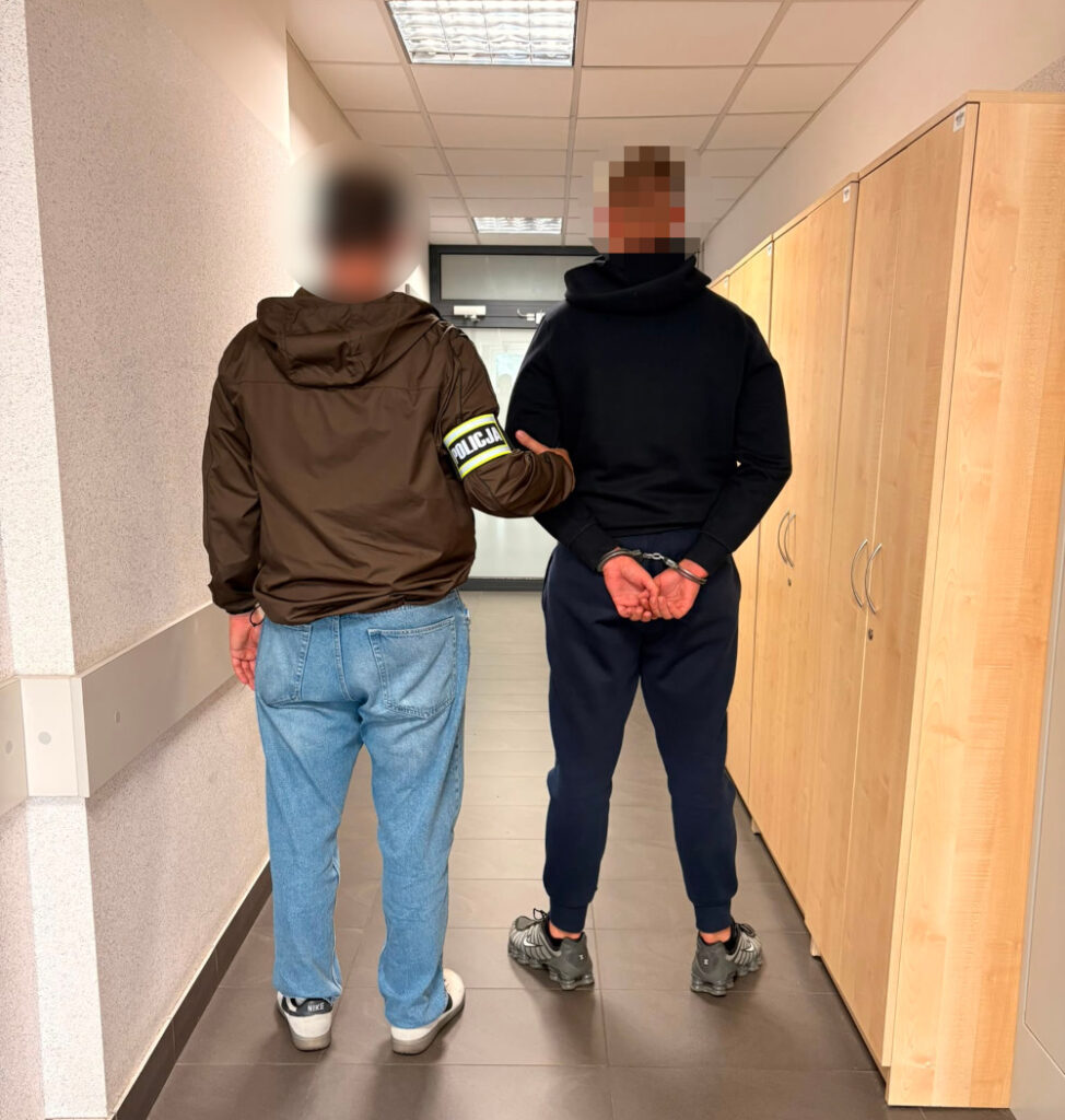 Policjant prowadzi zatrzymanego mężczyznę w kajdankach korytarzem w budynku administracyjnym.