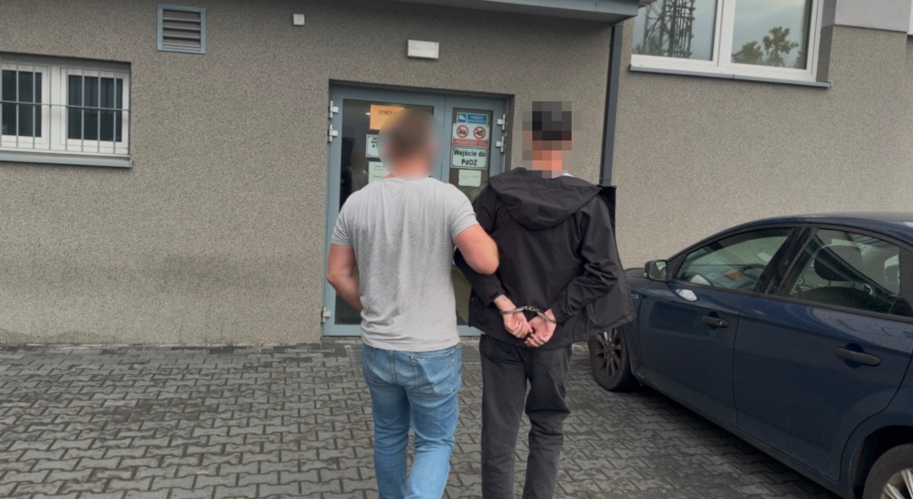 Mężczyzna w szarej koszulce prowadzi zatrzymanego mężczyznę w czarnej kurtce przed budynkiem z szarymi ścianami.