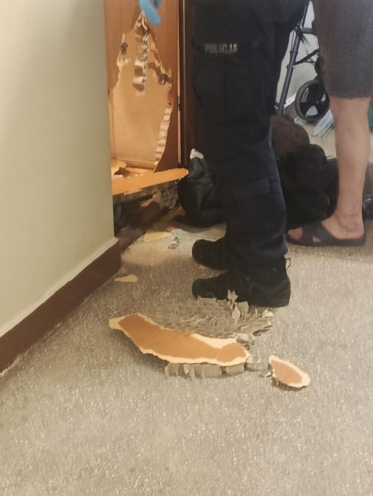 Policjant w mundurze z niebieskimi rękawiczkami stoi obok uszkodzonych drzwi w mieszkaniu, na podłodze leżą fragmenty drzwi.