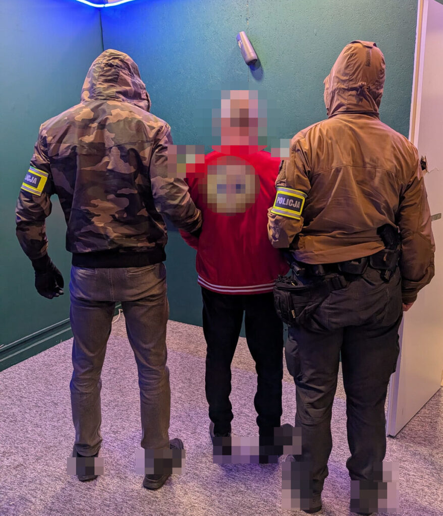 Mężczyźni w mundurach policyjnych zatrzymują osobę w czerwonej kurtce w pomieszczeniu z zieloną ścianą.