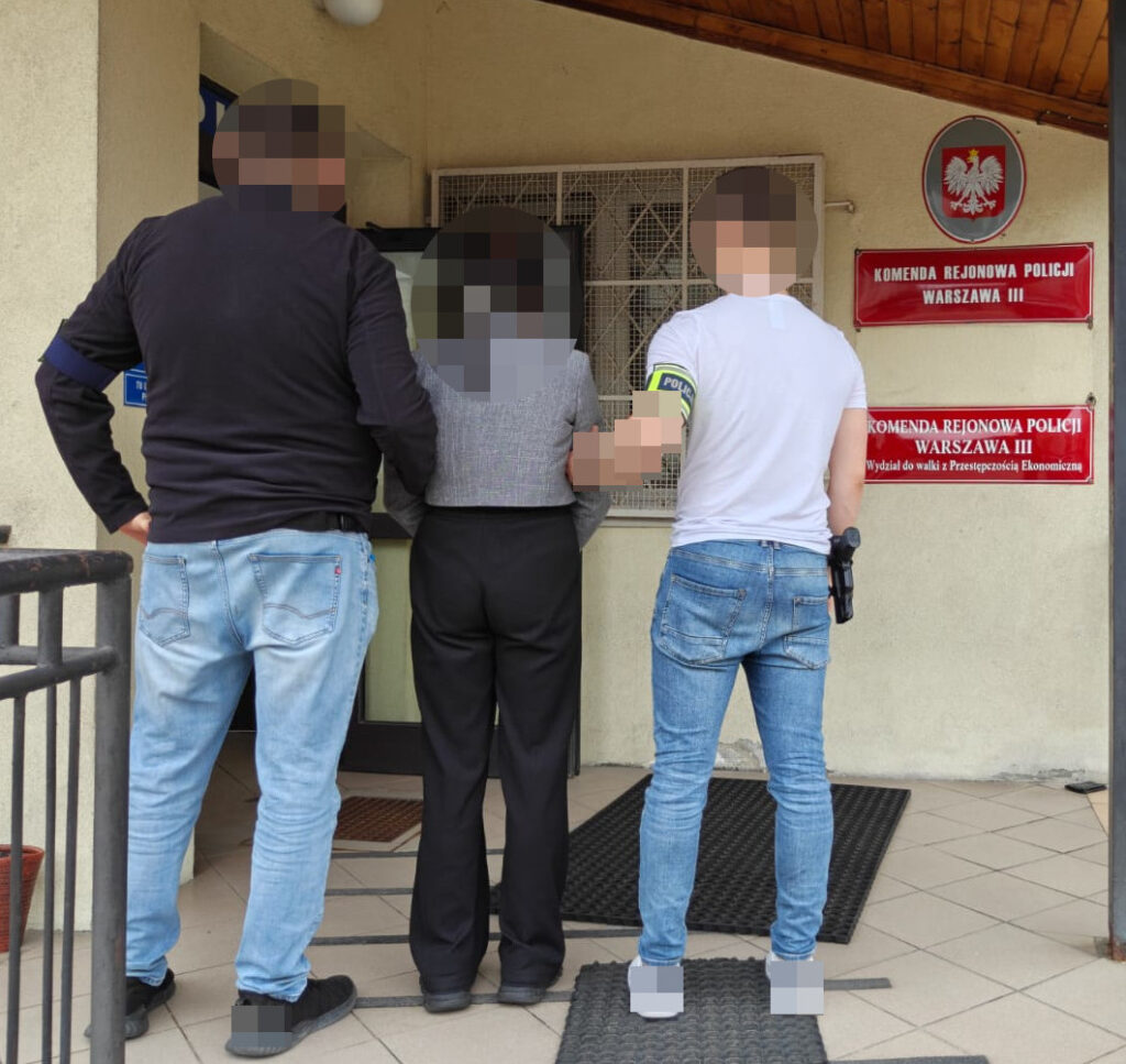 Dwie osoby zatrzymane przez policjantów przed budynkiem komendy rejonowej, w tle widoczny znak z nazwą jednostki.