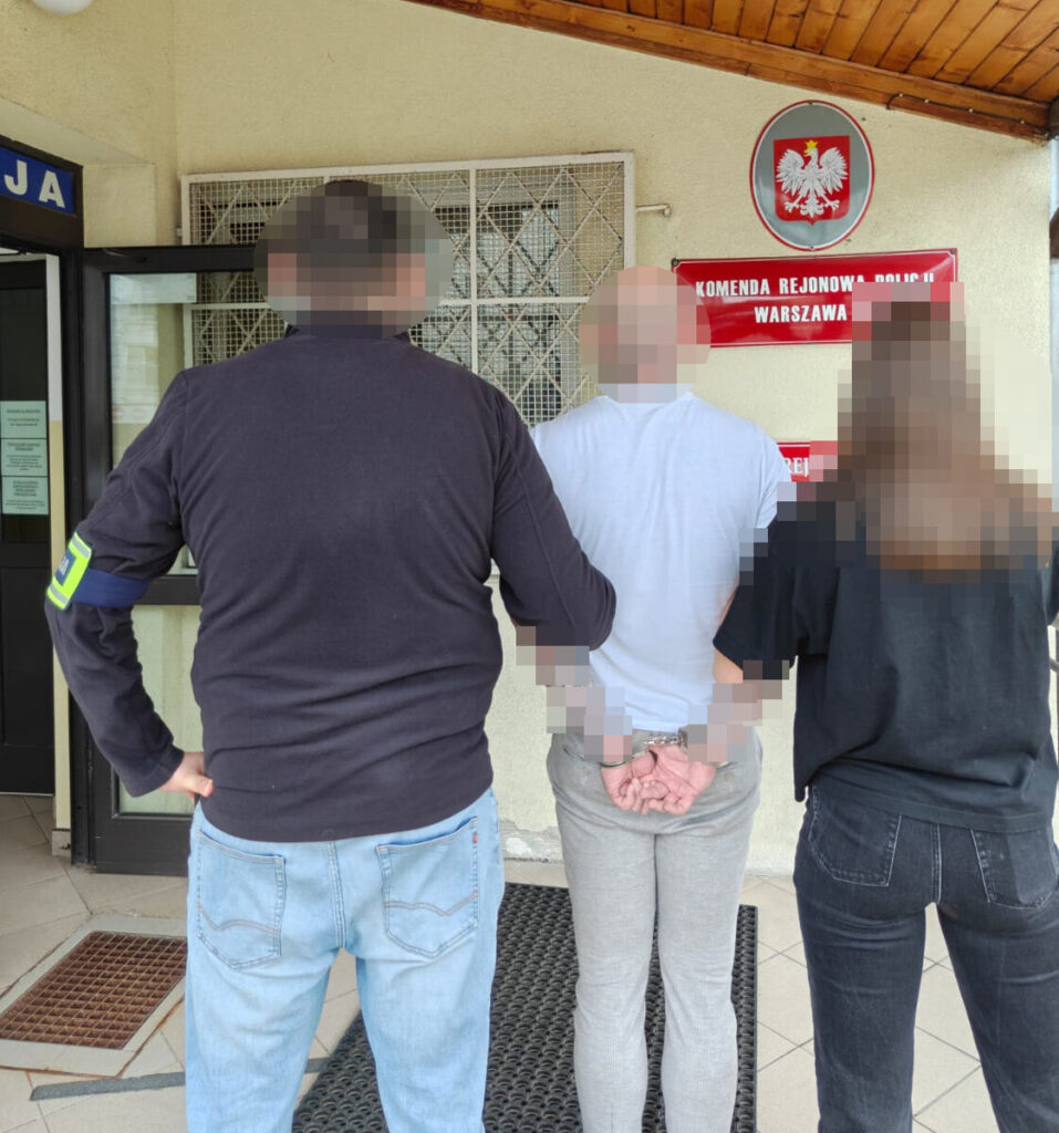 Mężczyzna w kajdankach stoi przed wejściem do budynku policji, a obok niego są dwie osoby, w tym funkcjonariusz.