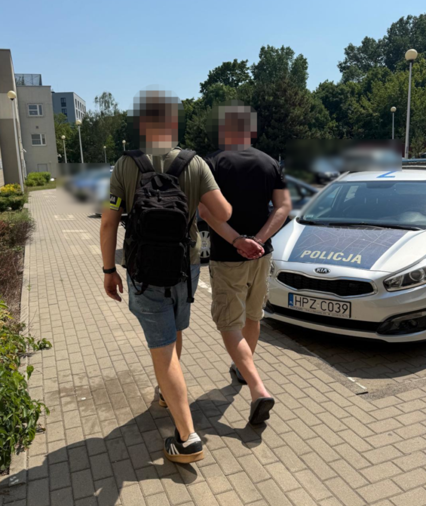 Mężczyzna w krótkich spodniach prowadzony przez policjanta w mundurze obok radiowozu w jasnym otoczeniu.